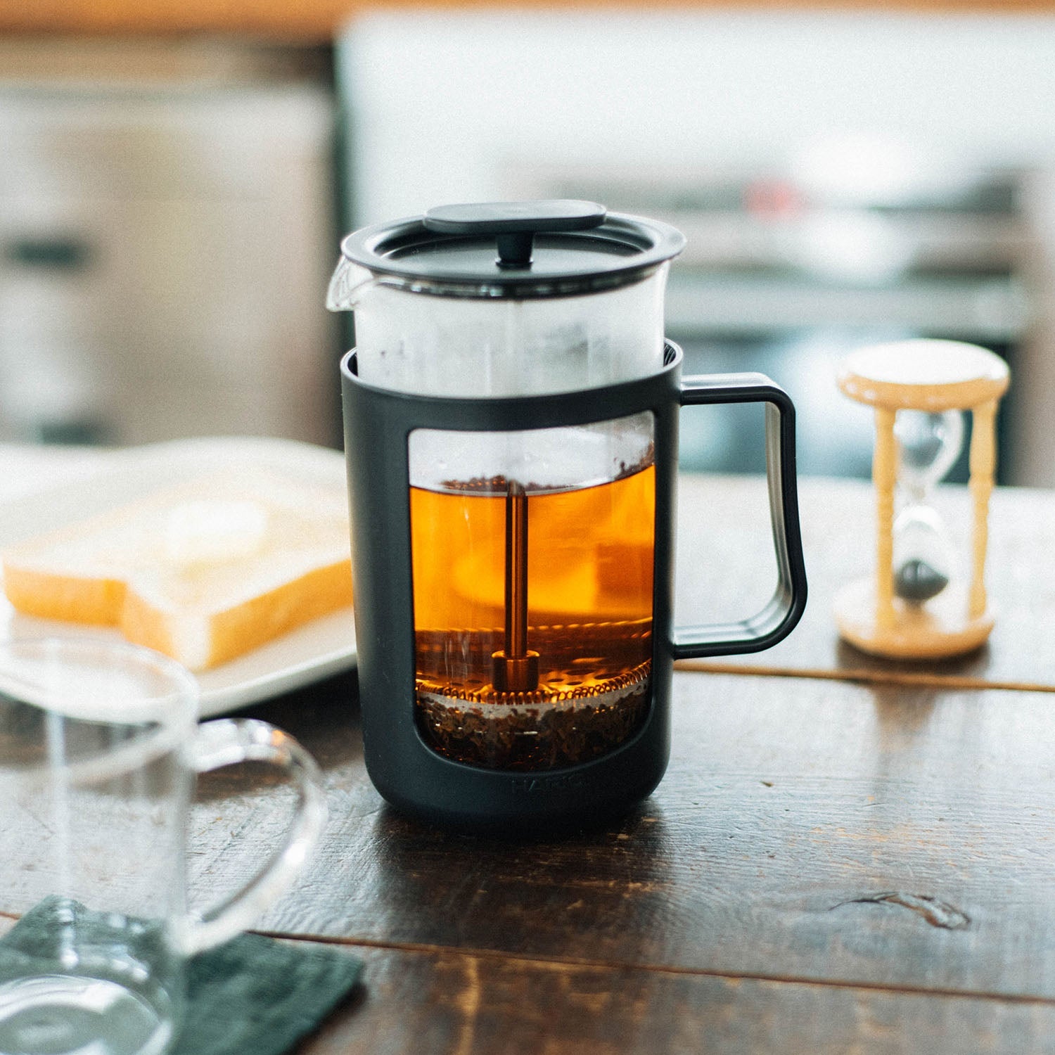Black tea and coffee maker with amber brewed beverage on wooden table with hourglass timer and toast nearby.