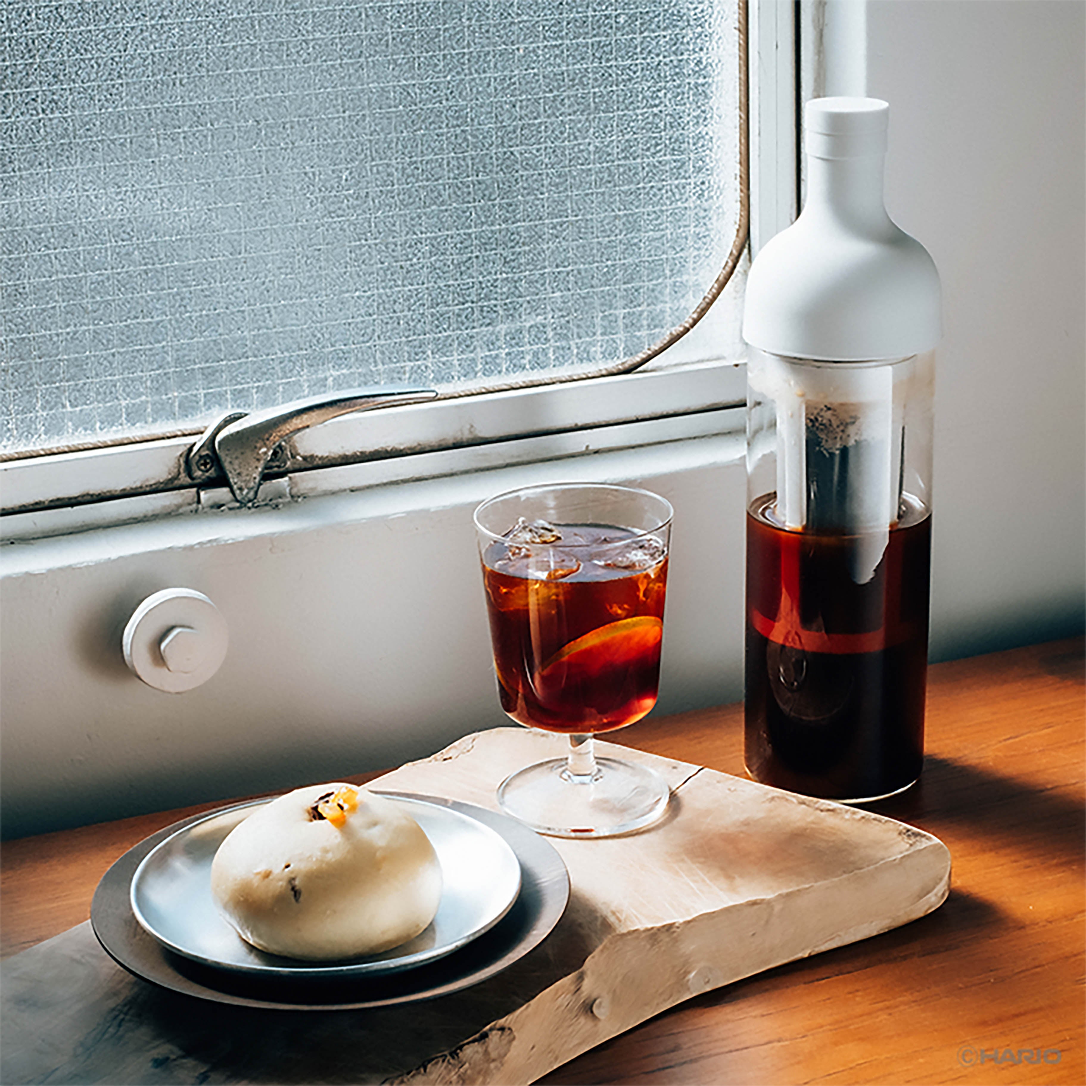 Cold brew coffee in glass bottle beside filled glass with ice cubes on wooden windowsill.