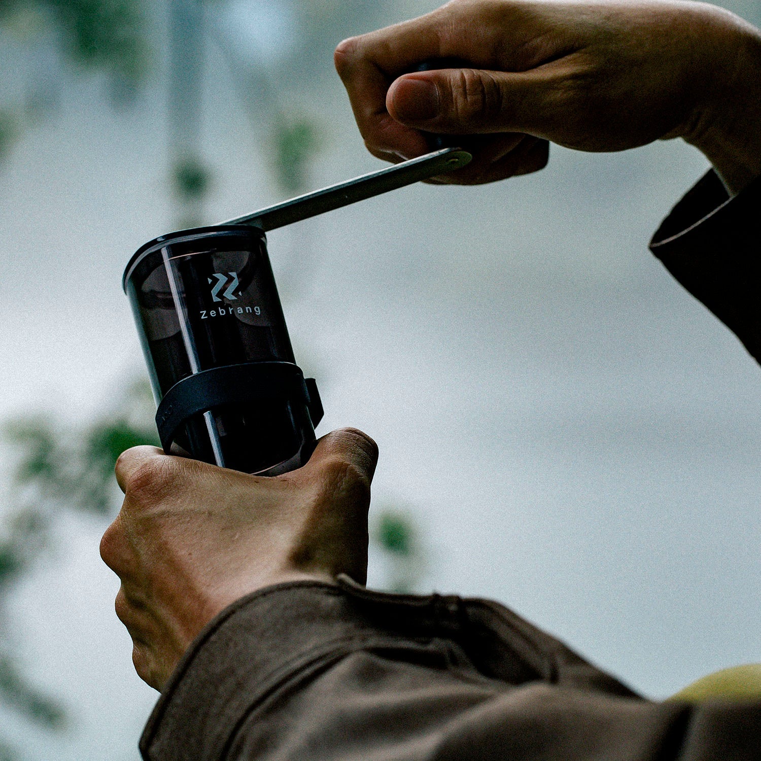 Hands holding a Zebrang outdoor coffee grinder with black body and logo visible against blurred natural background.