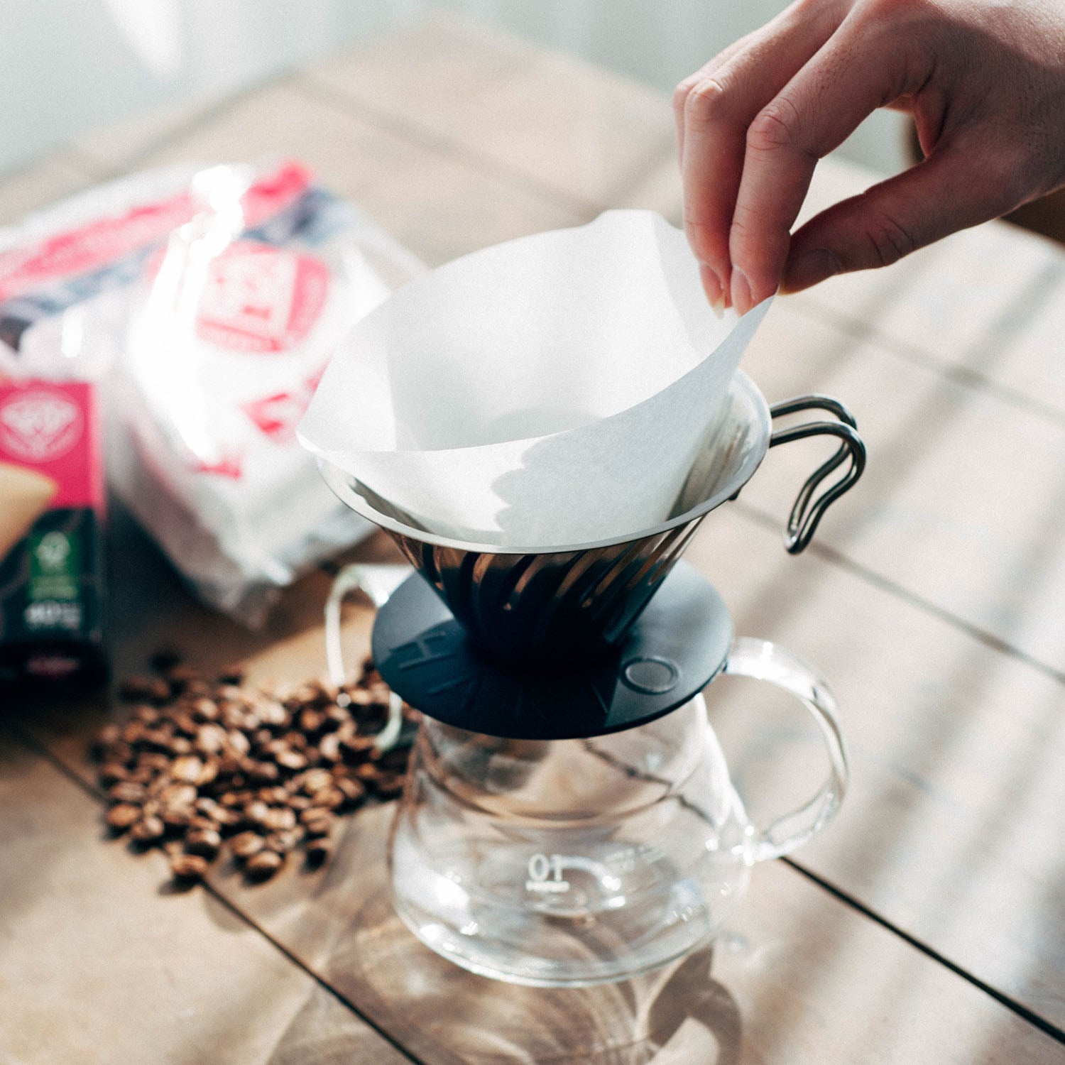 Hand placing white filter paper into black V60 dripper with glass server and coffee beans on wooden surface