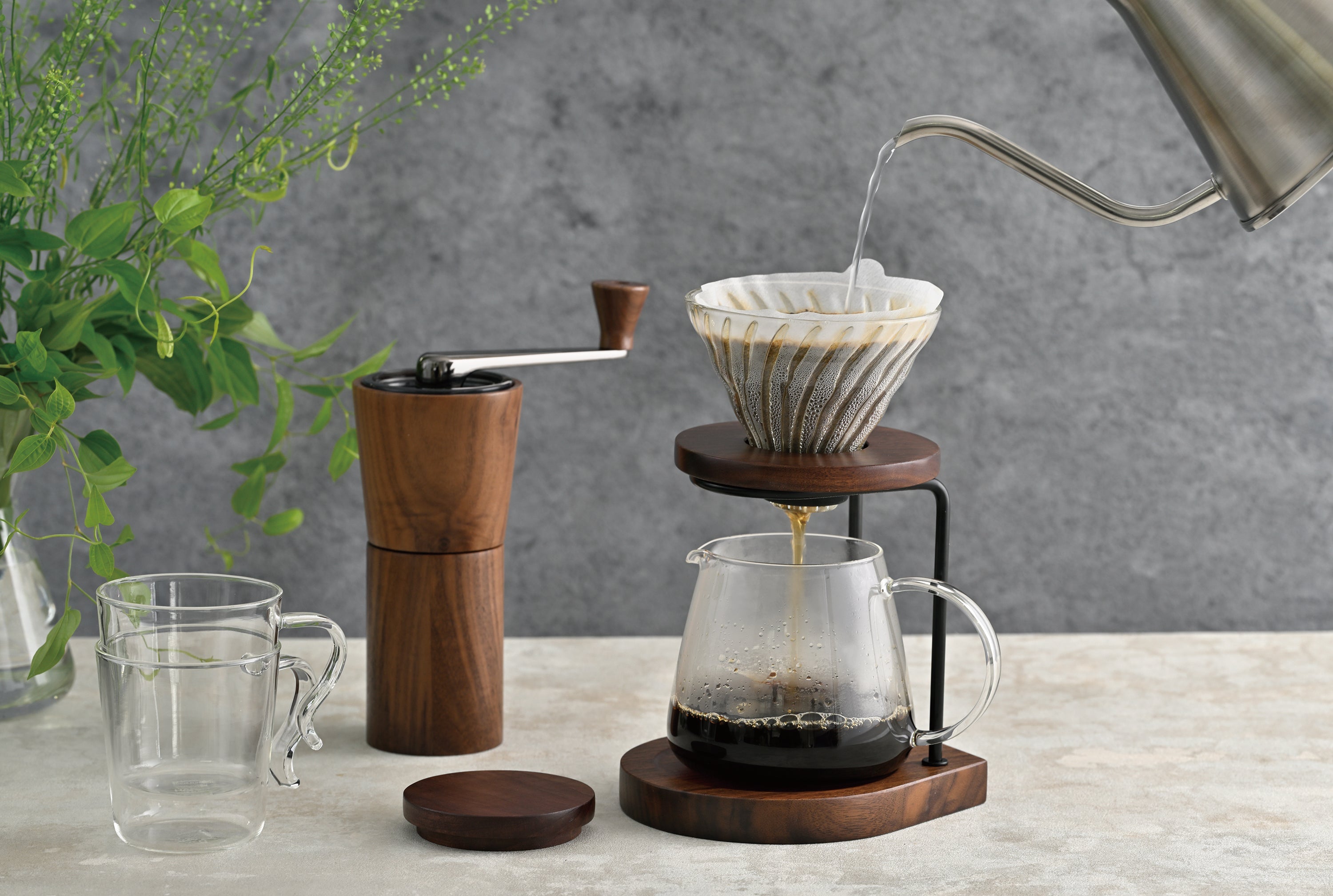Pour-over coffee setup with wooden grinder, ceramic dripper, glass server, and water being poured into filter.