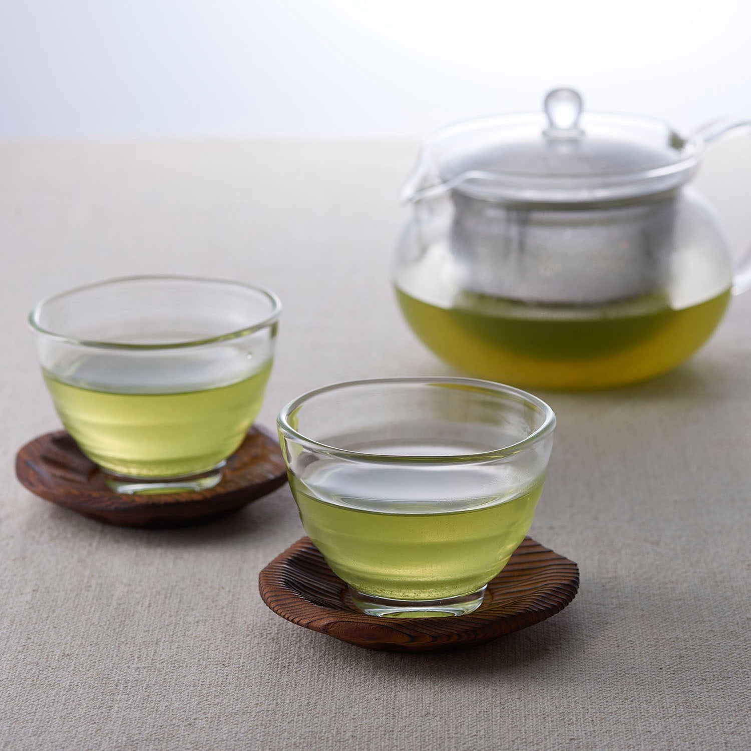 Two glass cups of green tea with wooden coasters and matching teapot with metal infuser.