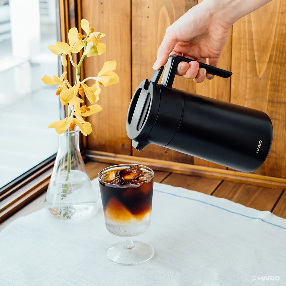 Hand pouring cold brew coffee from black ceramic-coated server into glass with ice beside yellow flowers and wooden backdrop.