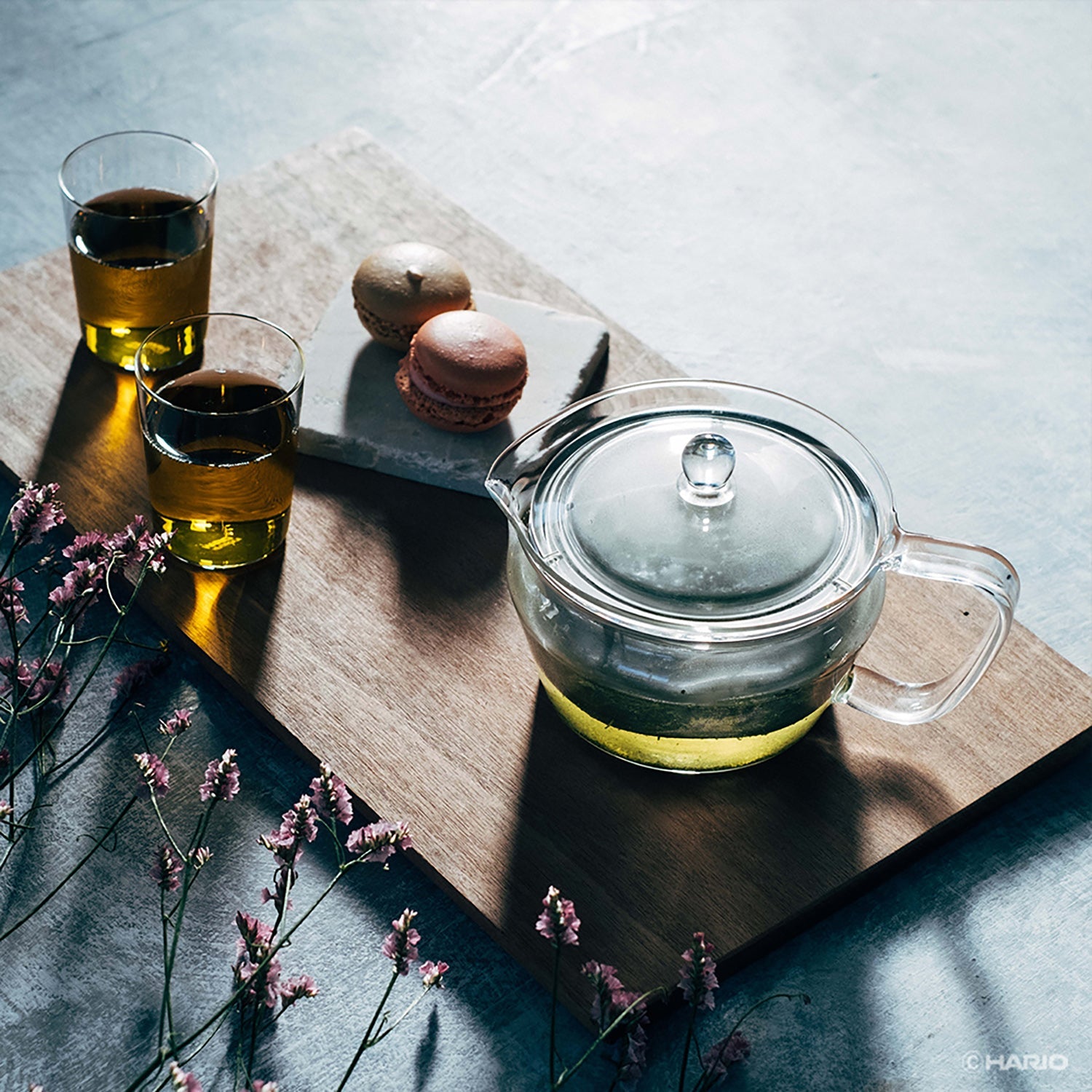 Glass tea pot with hot water beside two cups of tea, chocolate macarons, and dried flowers on wooden board.