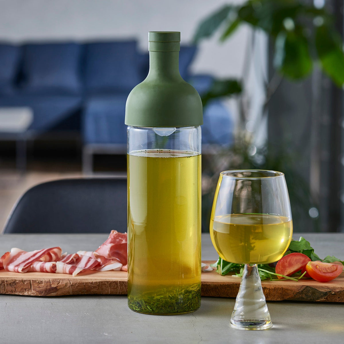 Olive-green glass cold brew pitcher with yellow infused tea, served in matching glass cup beside charcuterie board