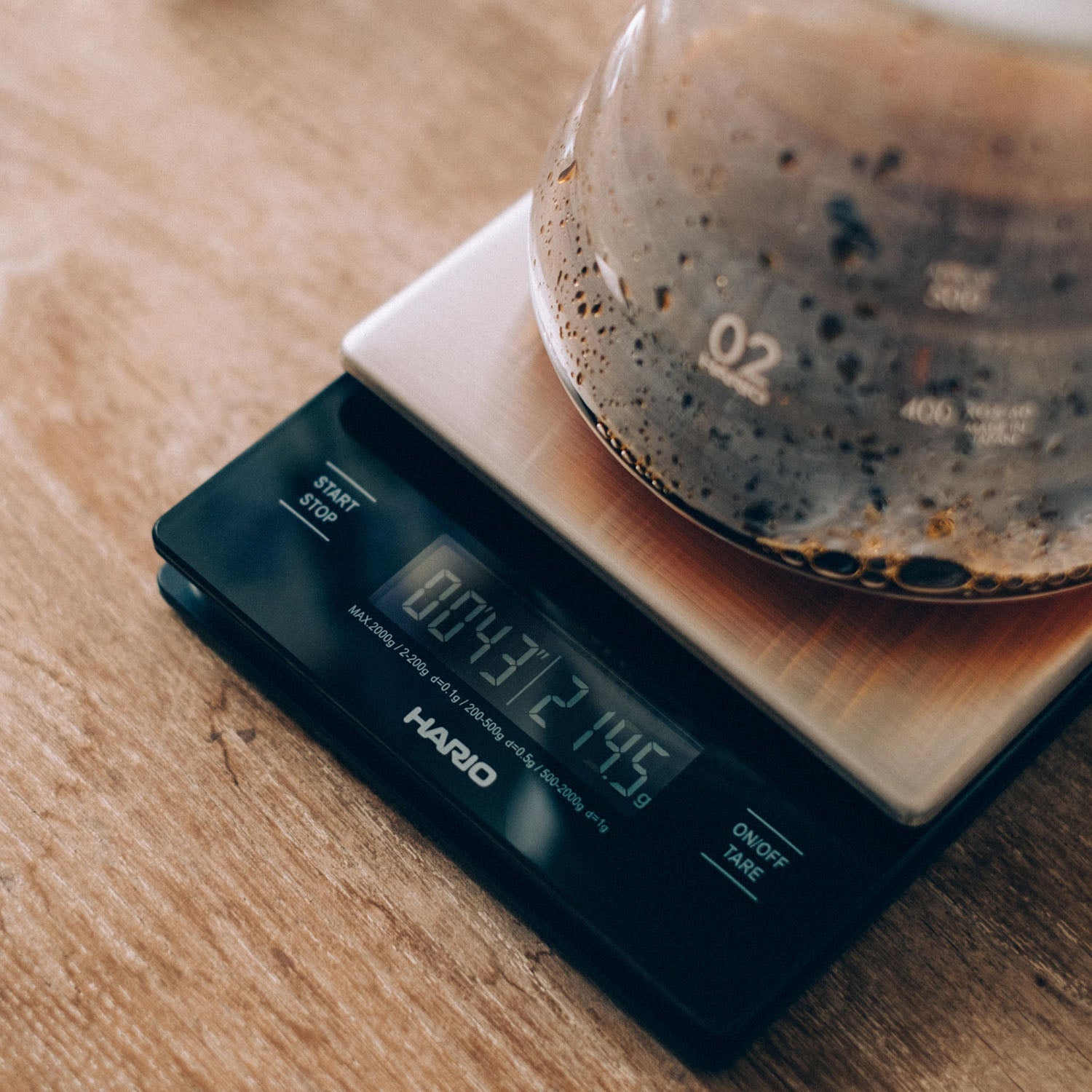 Digital kitchen scale with dark display showing measurement, pouring dark coffee into glass vessel on wooden surface.