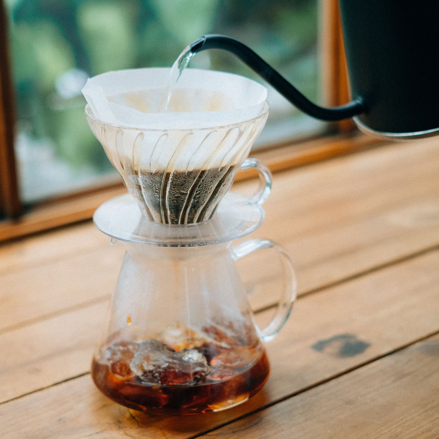 Hot water pouring into glass V60 dripper with coffee filter over glass carafe on wooden surface