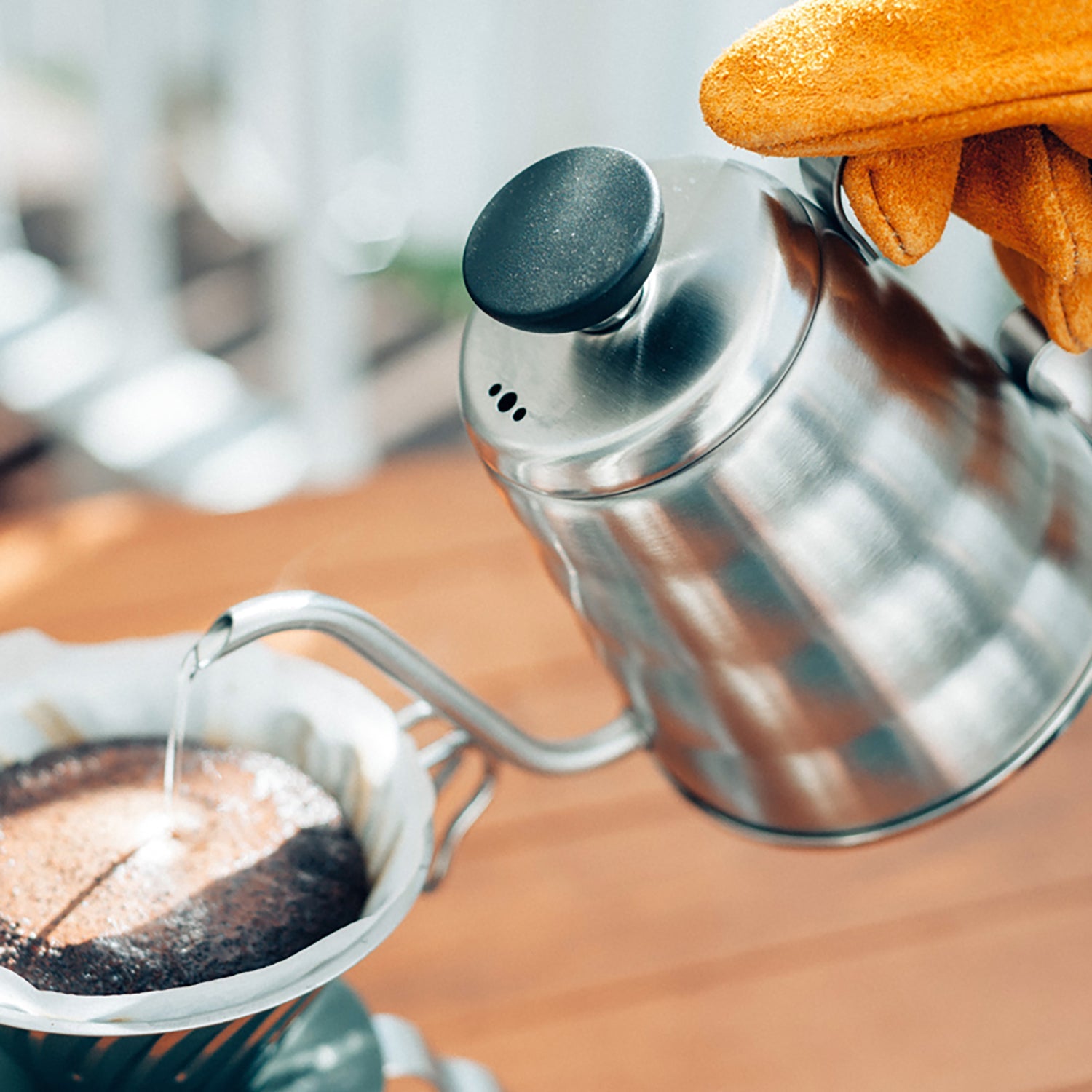 Stainless steel gooseneck kettle pouring water over coffee grounds with fresh bread nearby on wooden surface.