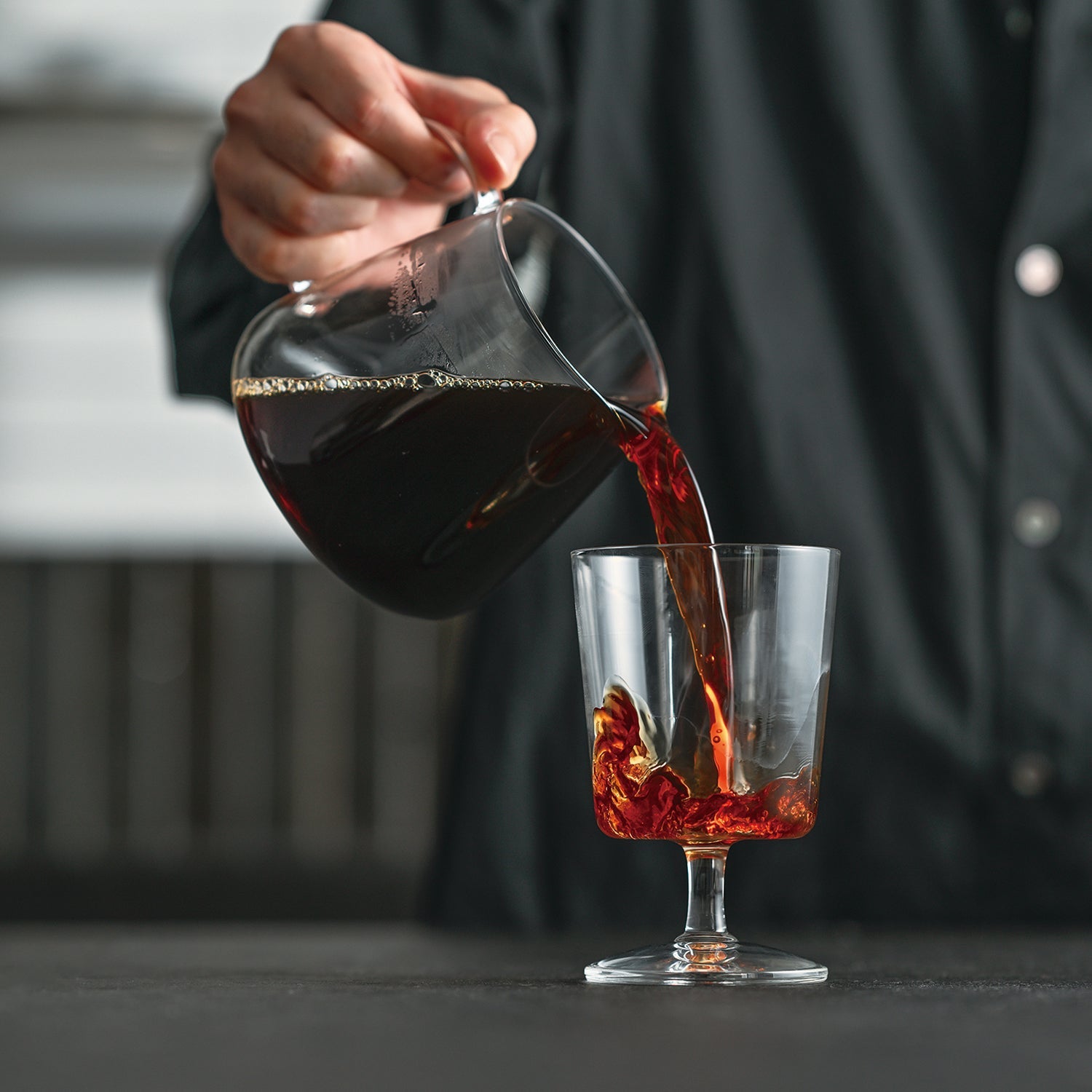 Person pouring dark red coffee from glass server into glass mug with ice