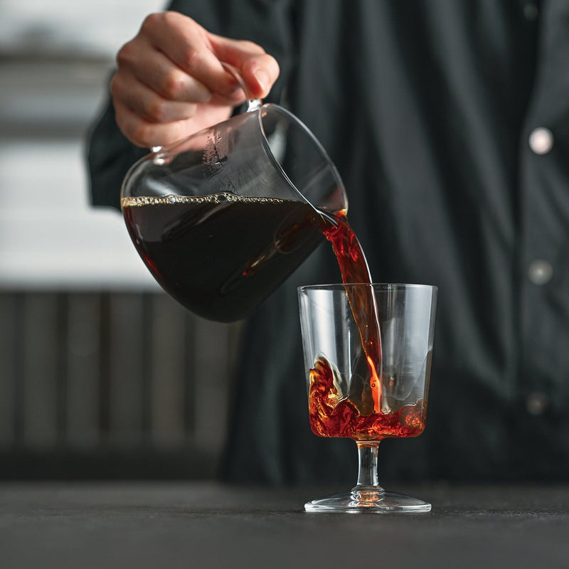 Person pouring dark red coffee from glass server into glass mug with ice
