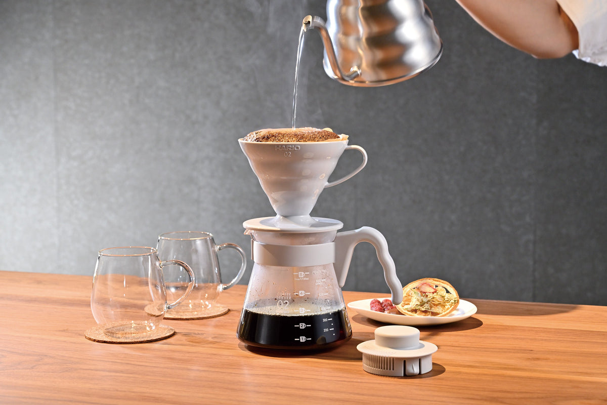 Pour-over coffee brewing in progress with hot water being poured into gray V60 dripper over glass carafe on wooden table.