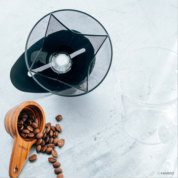 Overhead view of transparent Hario V60 dripper with wooden coffee scoop and roasted beans on surface