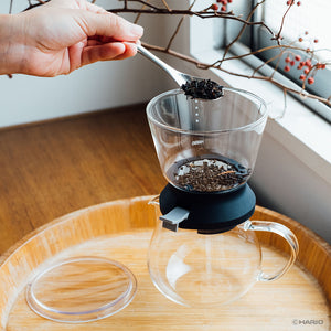Hands positioning Largo dripper above glass carafe to begin controlled tea steeping process.