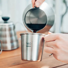 Hand pouring black coffee from silver Hario V60 stacking mug into matching silver camping cup on wooden surface