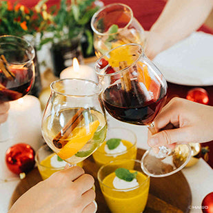 Multiple people holding tulip glasses filled with colorful beverages at holiday gathering with ornaments.