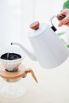 Person pouring hot water from white enamel kettle into coffee dripper with grounds
