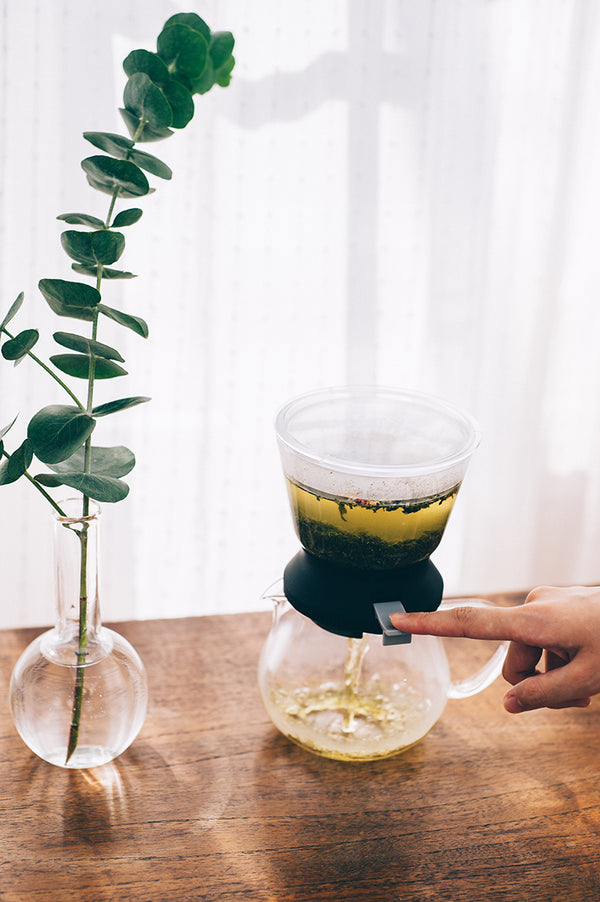 Hands releasing dark-brewed tea from Largo dripper into glass server below eucalyptus arrangement.