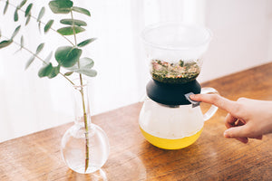 Hands adjusting Largo dripper's upper chamber containing loose tea leaves above yellow-filled server.