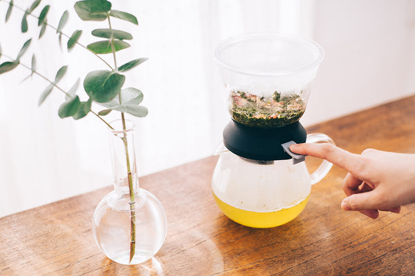 Hands adjusting Largo dripper's upper chamber containing loose tea leaves above yellow-filled server.