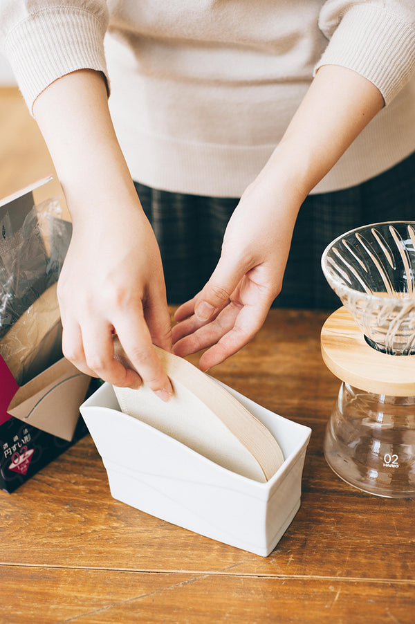 Hands placing unfolded coffee filters into white ceramic holder on wooden table