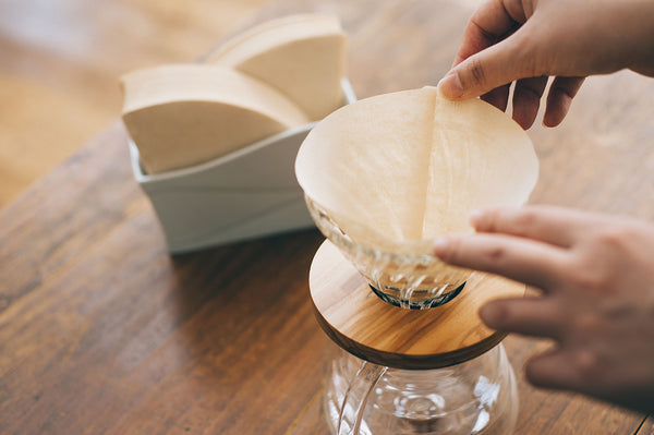 Hand inserting white V60 paper filter into glass dripper on wooden table.