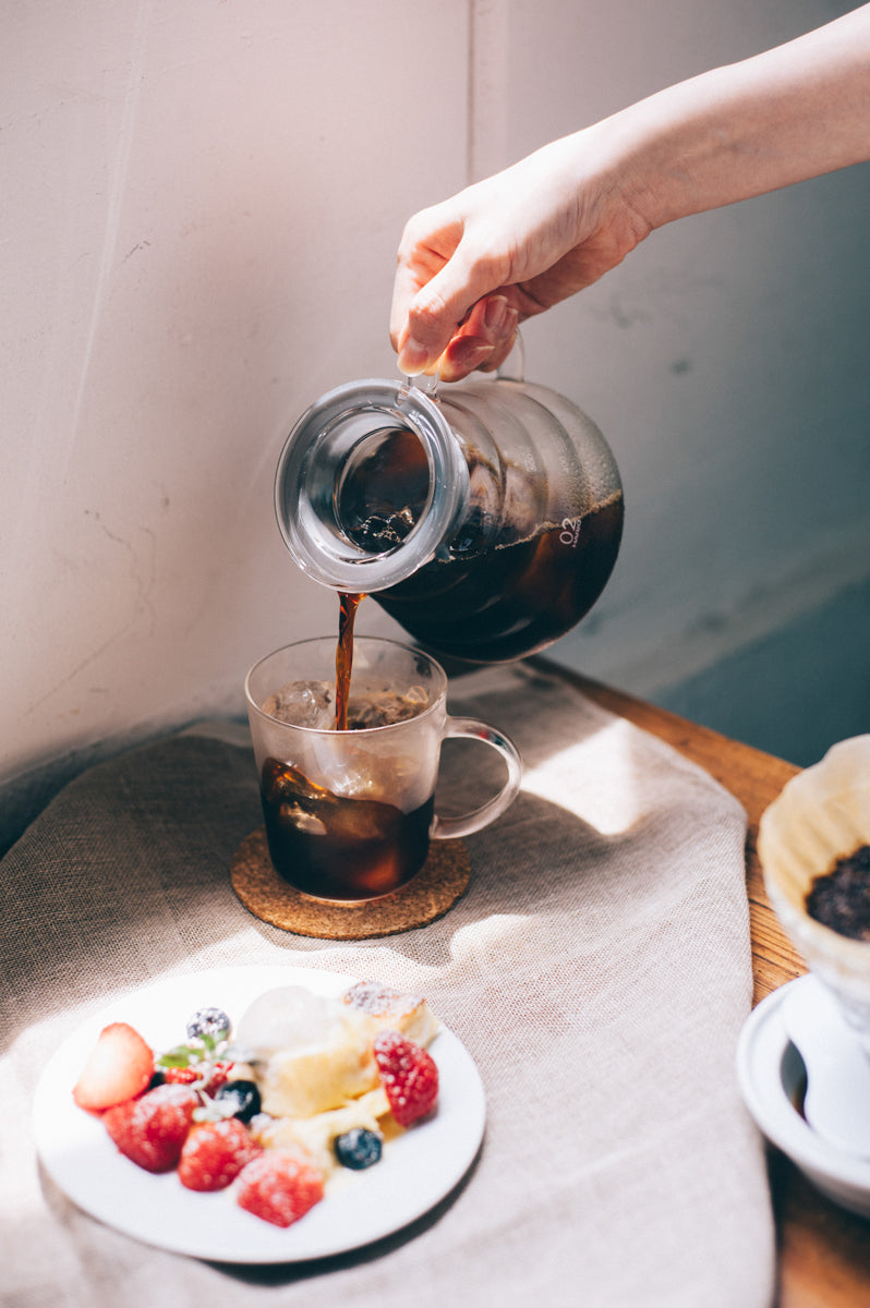Person pouring dark coffee from glass V60 server into clear mug with ice, surrounded by fresh berries and pastries on table.