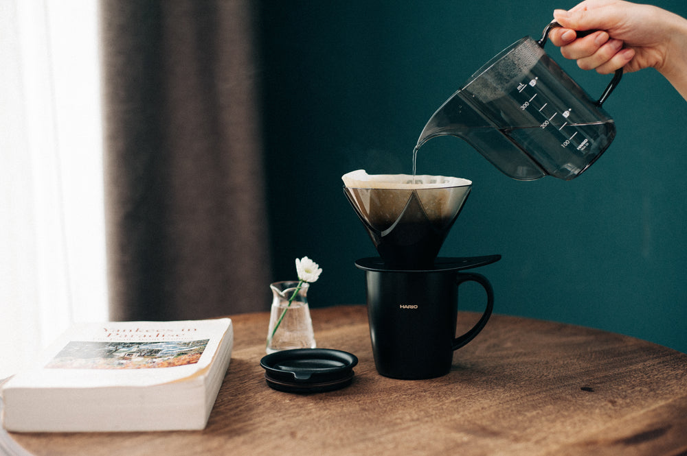 Person pouring hot water from black kettle into Hario V60 dripper over black mug on wooden table