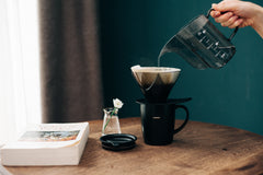 Person pouring hot water from black kettle into Hario V60 dripper over black mug on wooden table