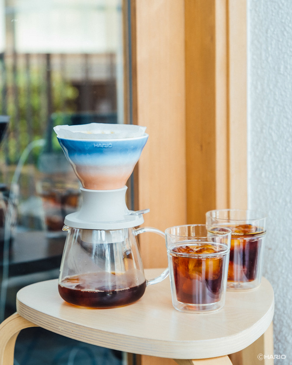 Man in blue shirt pouring water into white ceramic HARIO SWITCH dripper with gradient-striped filter cone on wooden counter