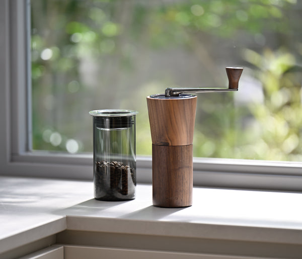 Walnut wood coffee grinder displayed on windowsill next to glass container of coffee beans.