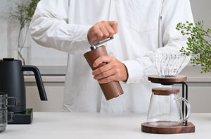 Hands operating walnut wood coffee grinder with glass carafe and pour-over equipment visible.