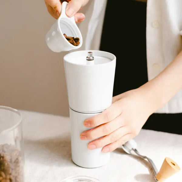 Hands pouring roasted coffee beans into white ceramic grinder's open top chamber.