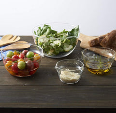 Glass mixing bowls with spouts surrounded by fresh salad ingredients, tomatoes, olive oil, and bread on dark wooden surface.