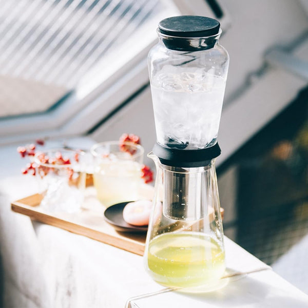 Two-chamber Shizuku dripper filled with water above and pale yellow brewed coffee below, styled on bright windowsill.