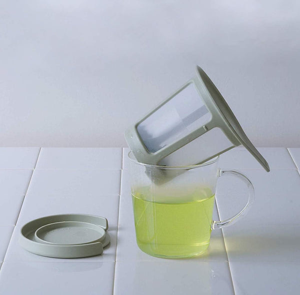 Tea infuser being lifted from mug of yellow-green tea, showing the mesh filter basket and ceramic lid resting on rim.