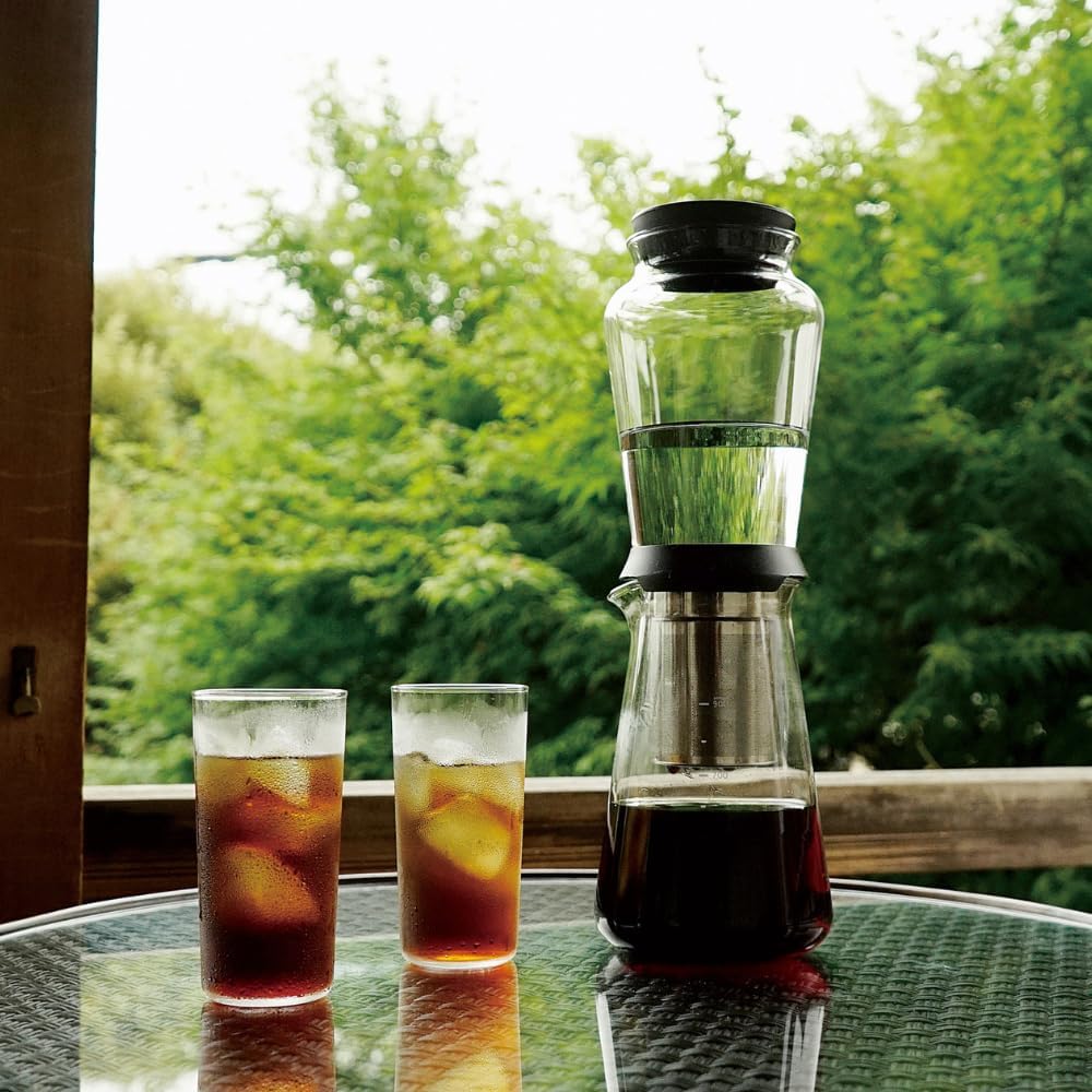 Shizuku cold brew coffee maker with two glasses of iced coffee beside it, overlooking green foliage through a window.