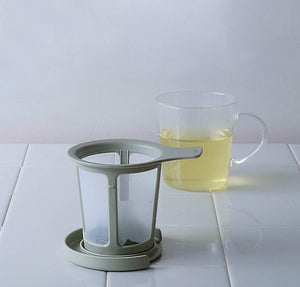 Smoky green one-cup tea maker with mesh infuser positioned next to glass mug filled with pale yellow tea.
