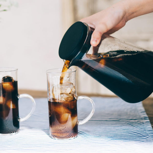 Hand pouring dark brewed coffee from Shizuku carafe into glass filled with ice and coffee.