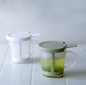 Two one-cup tea makers displayed side by side: white ceramic and smoky green, both with glass mugs and lids.