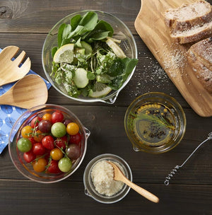 Overhead view of glass mixing bowl with spout containing leafy greens and sliced pears, with tomatoes, olive oil, flour