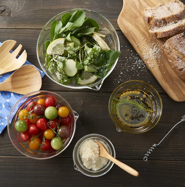 Overhead view of glass mixing bowl with spout containing leafy greens and sliced pears, with tomatoes, olive oil, flour
