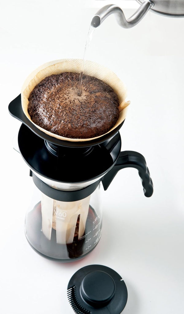 Hot water brewing dark coffee grounds in paper filter inside black dripper basket with ice strainer below.