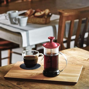 Red Hario Cafepress with black plunger filled with brewed coffee, served in glass cup on wooden tray.