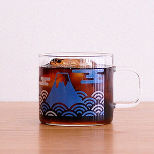 Glass mug filled with dark beverage, displaying Mount Fuji design in blue with pink clouds.