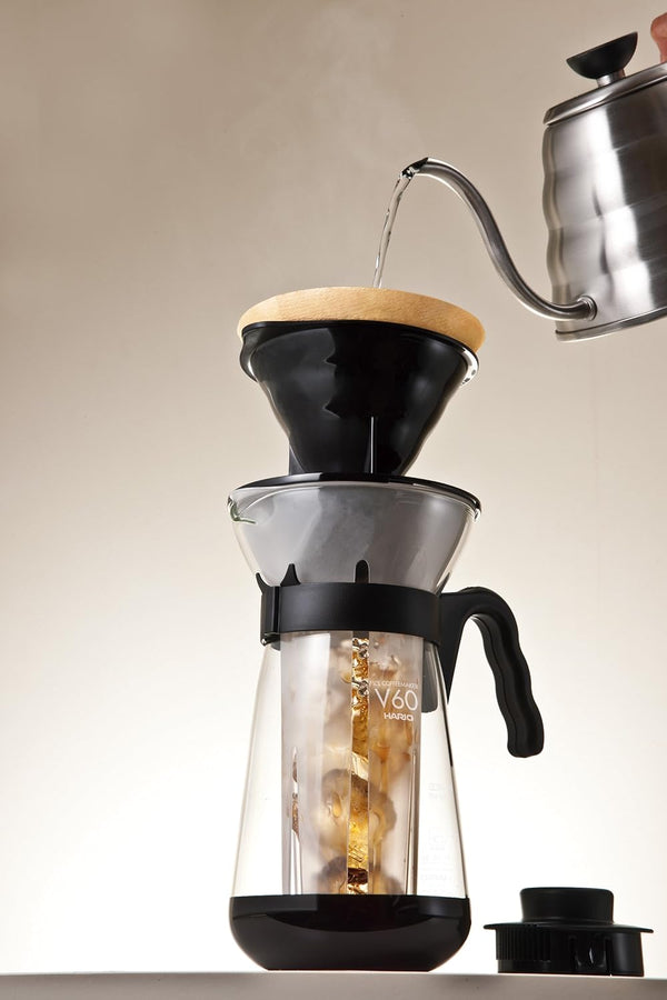 Hario V60 coffee maker with water pouring from kettle into black dripper basket with wooden rim.