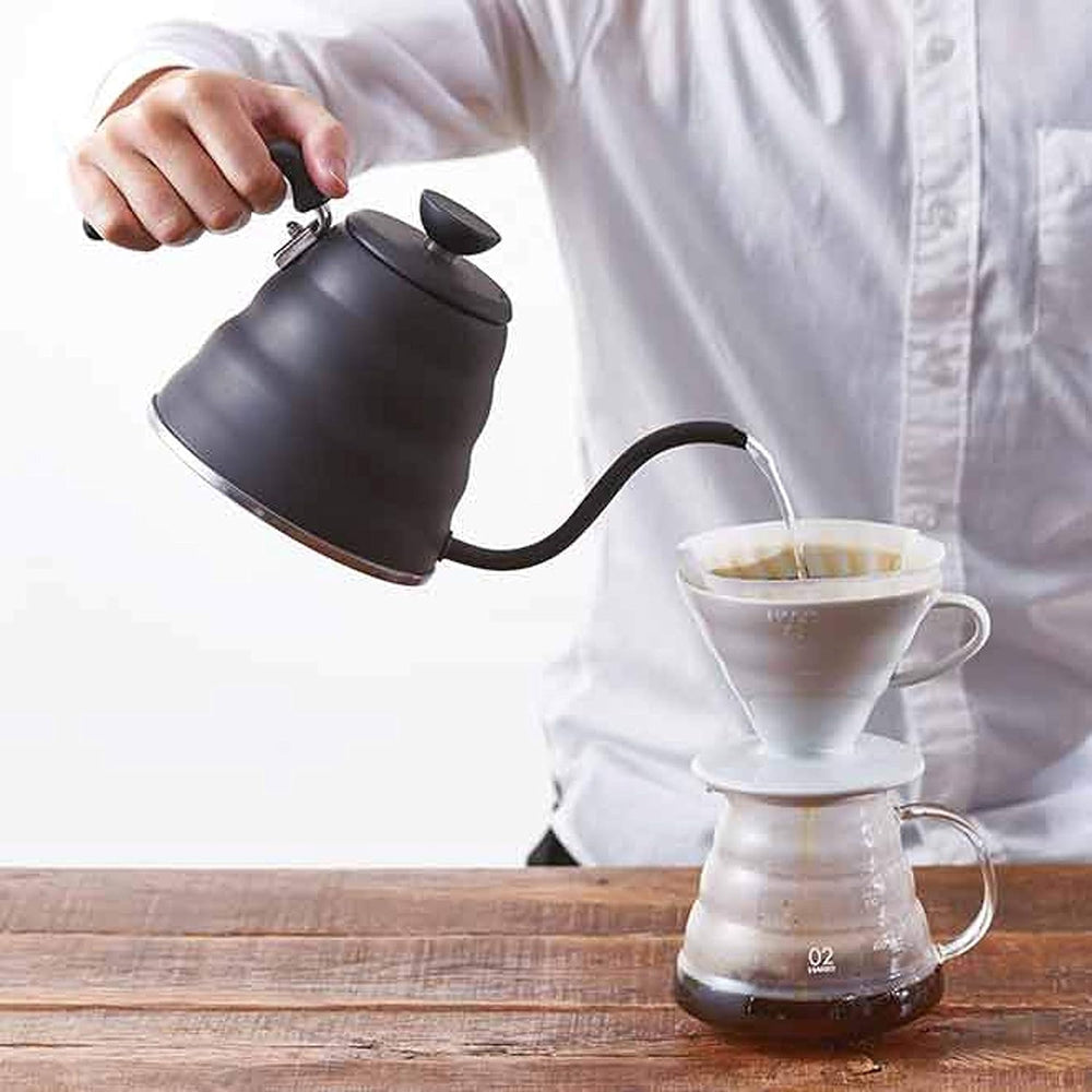 Person pouring hot water from black gooseneck kettle into white coffee dripper positioned over glass carafe on wooden table.