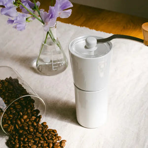 White ceramic coffee grinder with glass burr chamber displayed among purple flowers and roasted coffee beans.