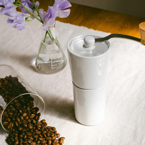 White ceramic coffee grinder with glass burr chamber displayed among purple flowers and roasted coffee beans.
