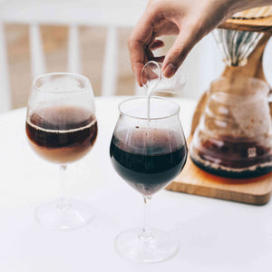 Person pouring dark coffee into tulip glass beside another filled glass and brewing equipment.