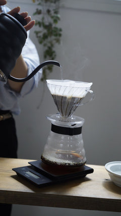 Person pouring hot water from gooseneck kettle into V60 dripper with white filter over black digital scale on wooden table